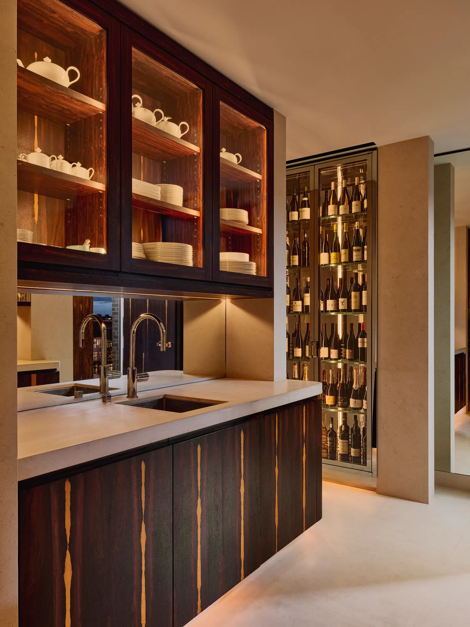 Contemporary kitchenette with dark wood cabinetry, open shelves displaying white crockery, and a marble countertop with twin sinks, adjacent to a glass-fronted wine display softly lit with warm ambient lighting.
