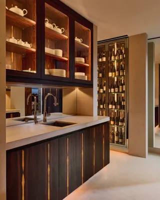 Contemporary kitchenette with dark wood cabinetry, open shelves displaying white crockery, and a marble countertop with twin sinks, adjacent to a glass-fronted wine display softly lit with warm ambient lighting.
