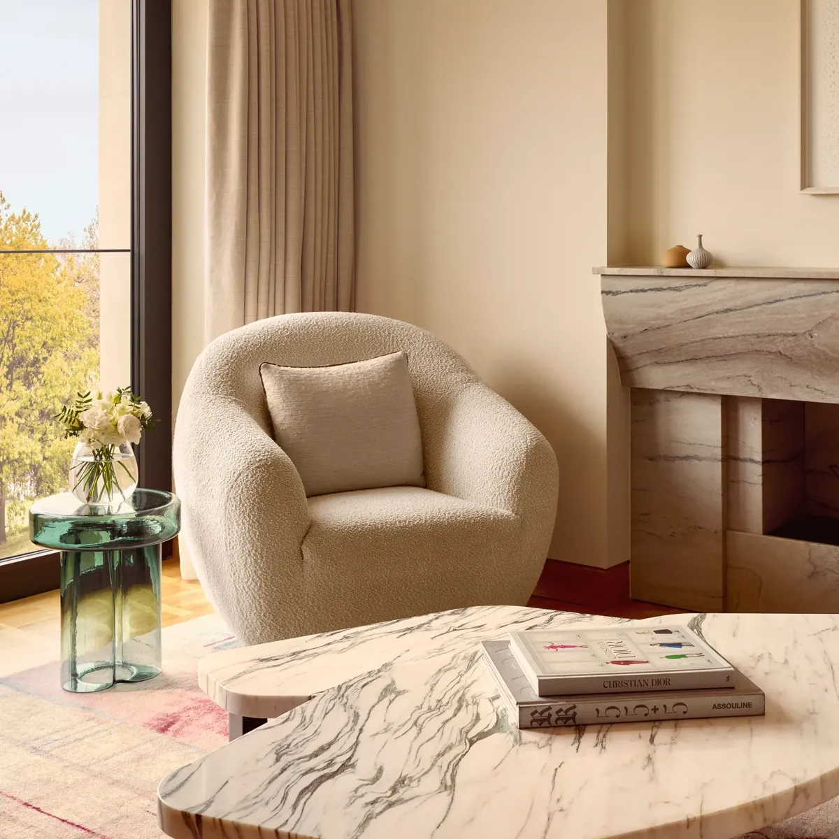 Cozy seating corner featuring a boucle armchair, green glass side table with flowers, and marble coffee table, set beside floor-to-ceiling windows.