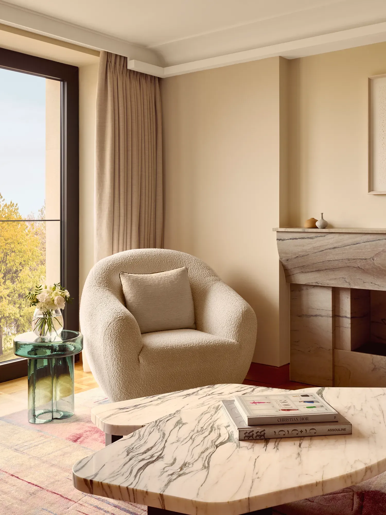 Cozy seating corner featuring a boucle armchair, green glass side table with flowers, and marble coffee table, set beside floor-to-ceiling windows.