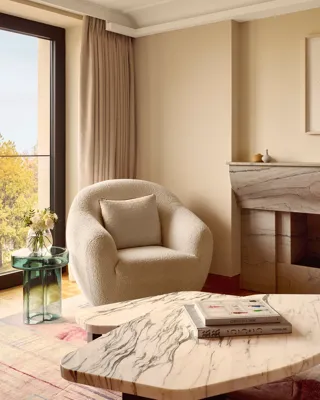 Cozy seating corner featuring a boucle armchair, green glass side table with flowers, and marble coffee table, set beside floor-to-ceiling windows.