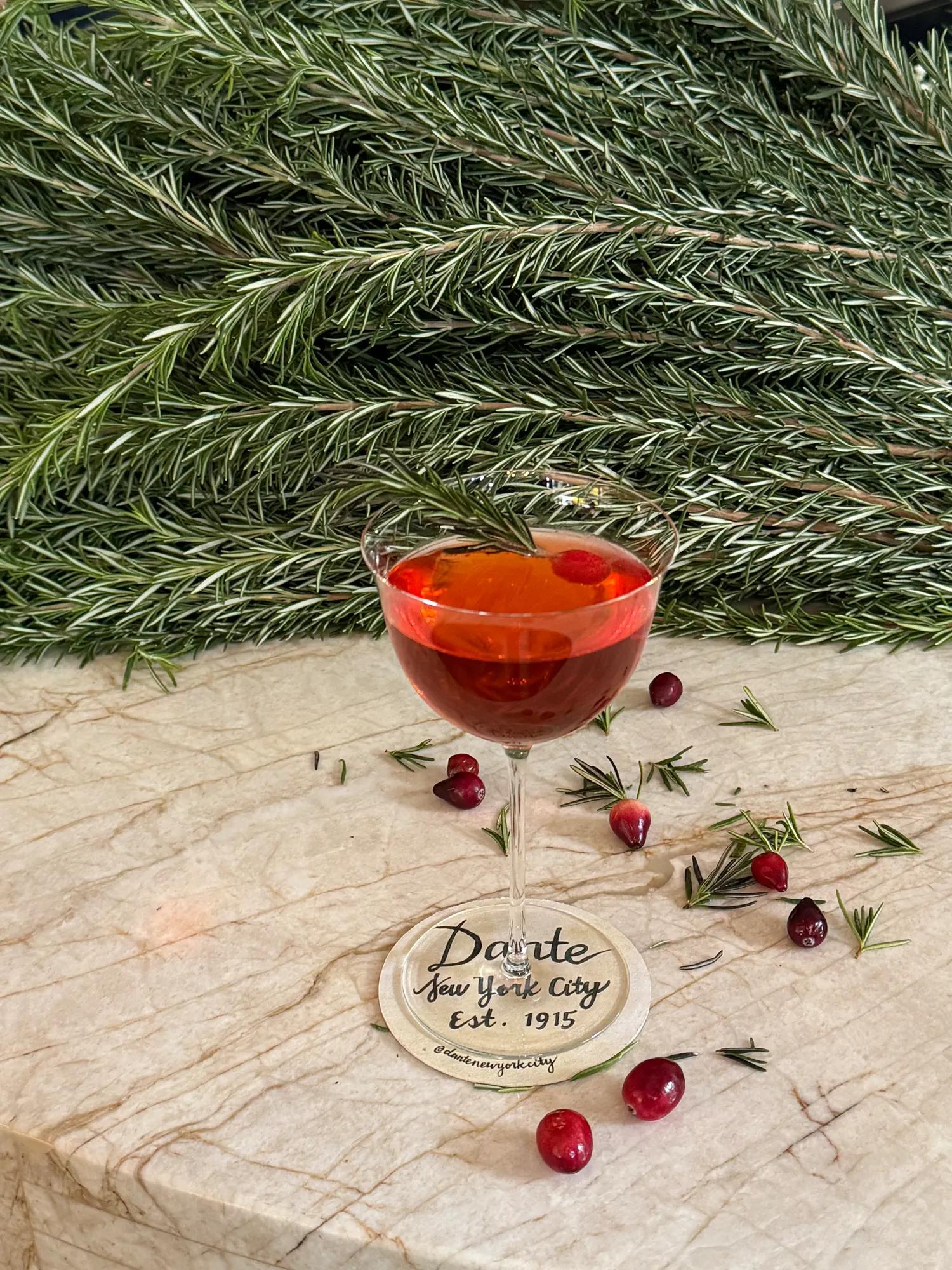A stemmed cocktail glass filled with a deep ruby-red cocktail sits on a light marble surface, garnished with a fresh rosemary sprig. Scattered cranberries and rosemary sprigs surround the glass, while a lush backdrop of evergreen branches creates a festive holiday setting. The glass rests on a coaster displaying the text “Dante New York City Est. 1915