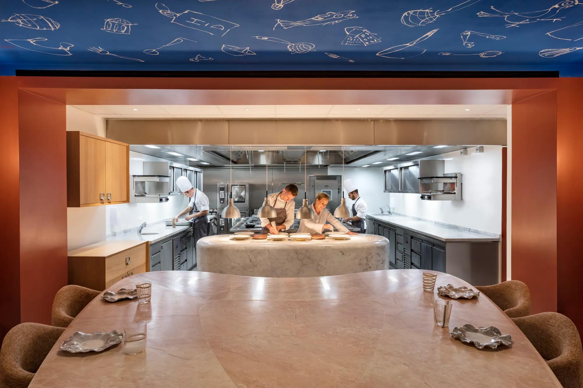Chefs prepare dishes in an open kitchen, viewed from a marble dining counter set with plates and glasses.