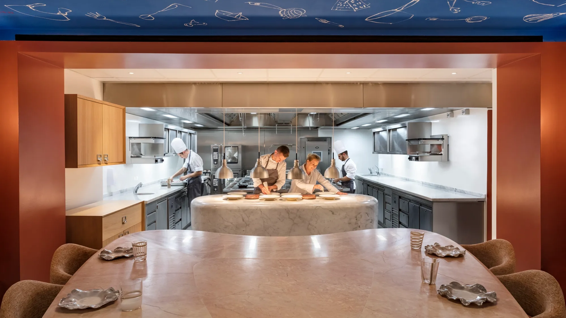Chefs prepare dishes in an open kitchen, viewed from a marble dining counter set with plates and glasses.