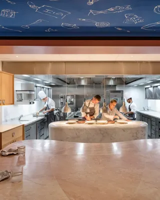 Chefs prepare dishes in an open kitchen, viewed from a marble dining counter set with plates and glasses.