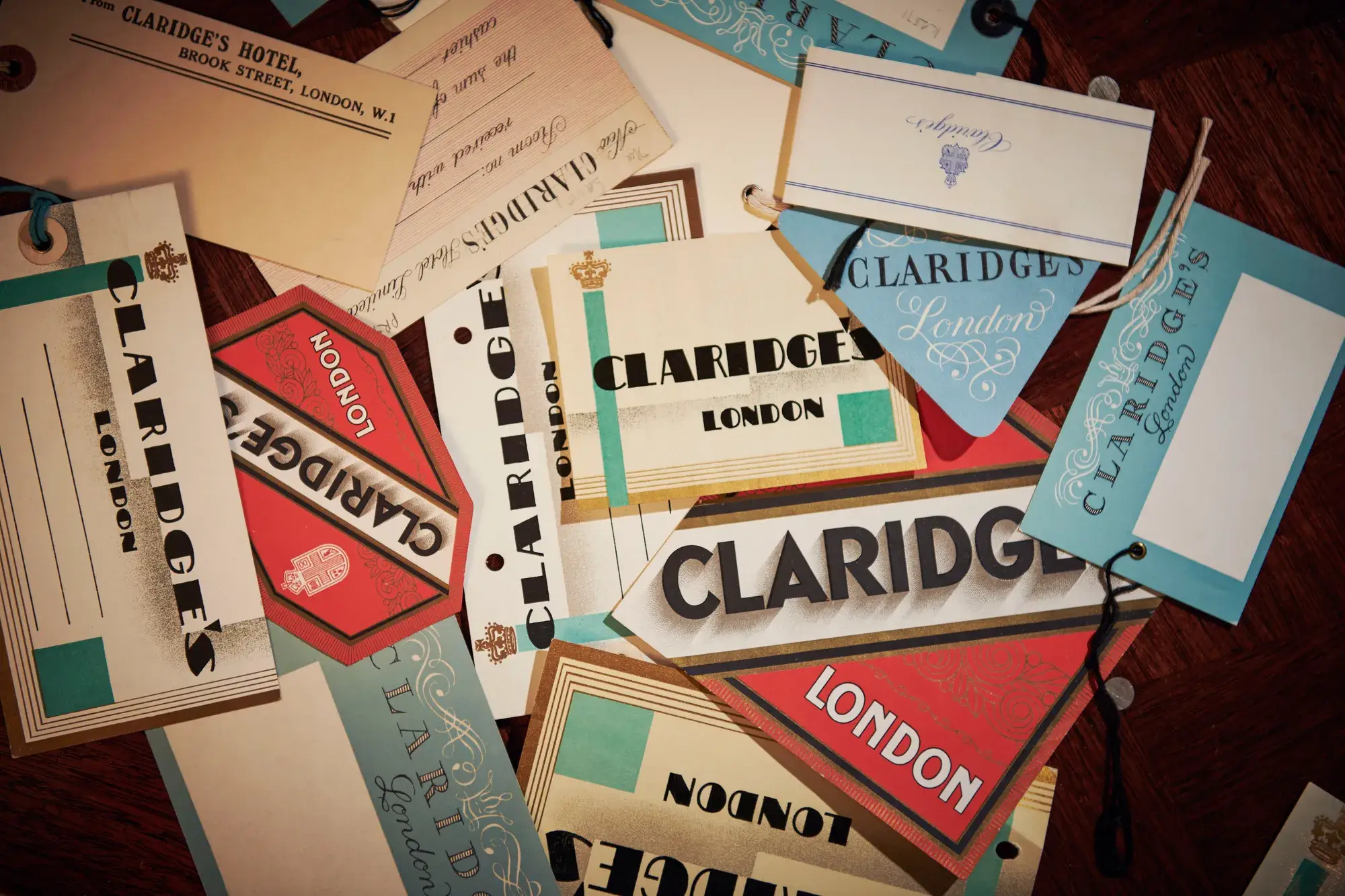 Collection of vintage Claridge’s luggage labels and tags scattered on a wooden surface in teal, cream, and red tones.