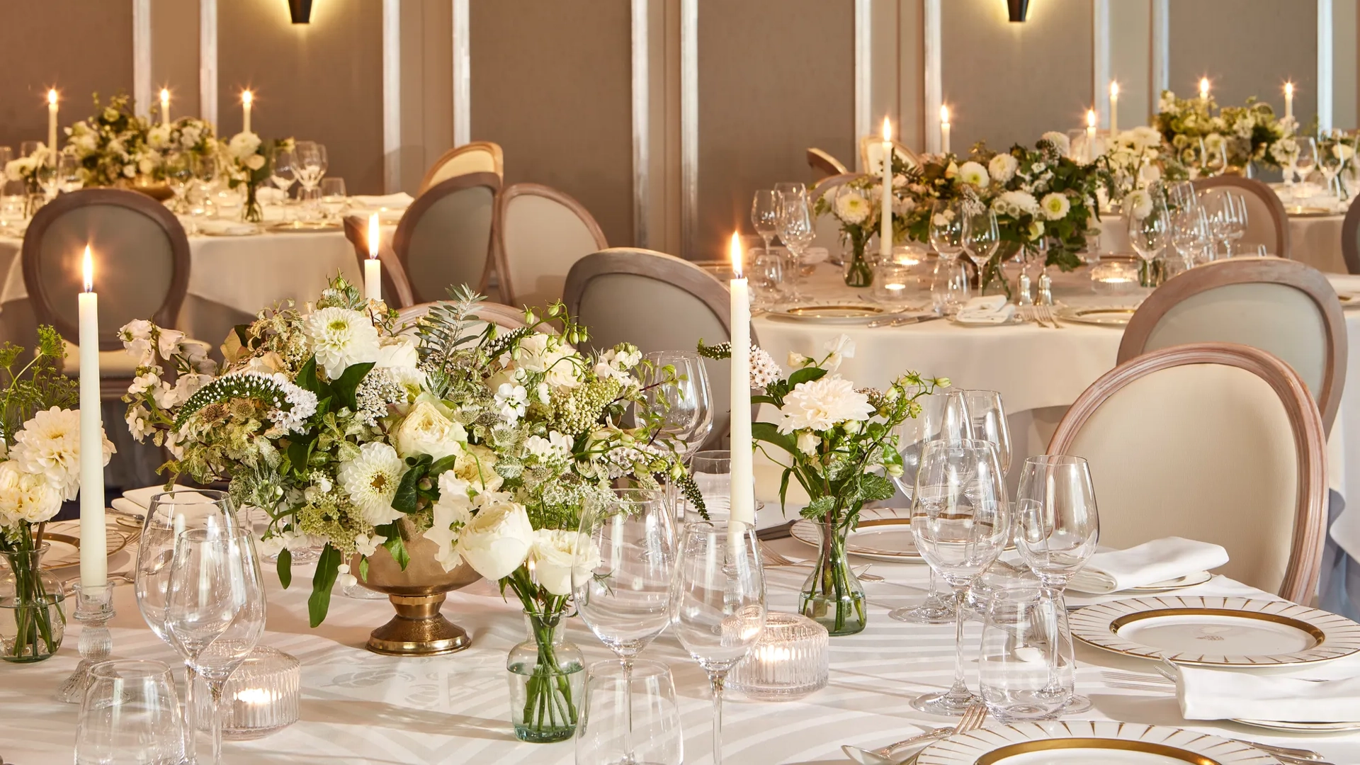 Elegant wedding reception setup in a softly lit dining room, featuring round tables dressed in white linen, gold-rimmed plates, crystal glassware, and floral centrepieces of white roses and greenery surrounded by tall lit candles.