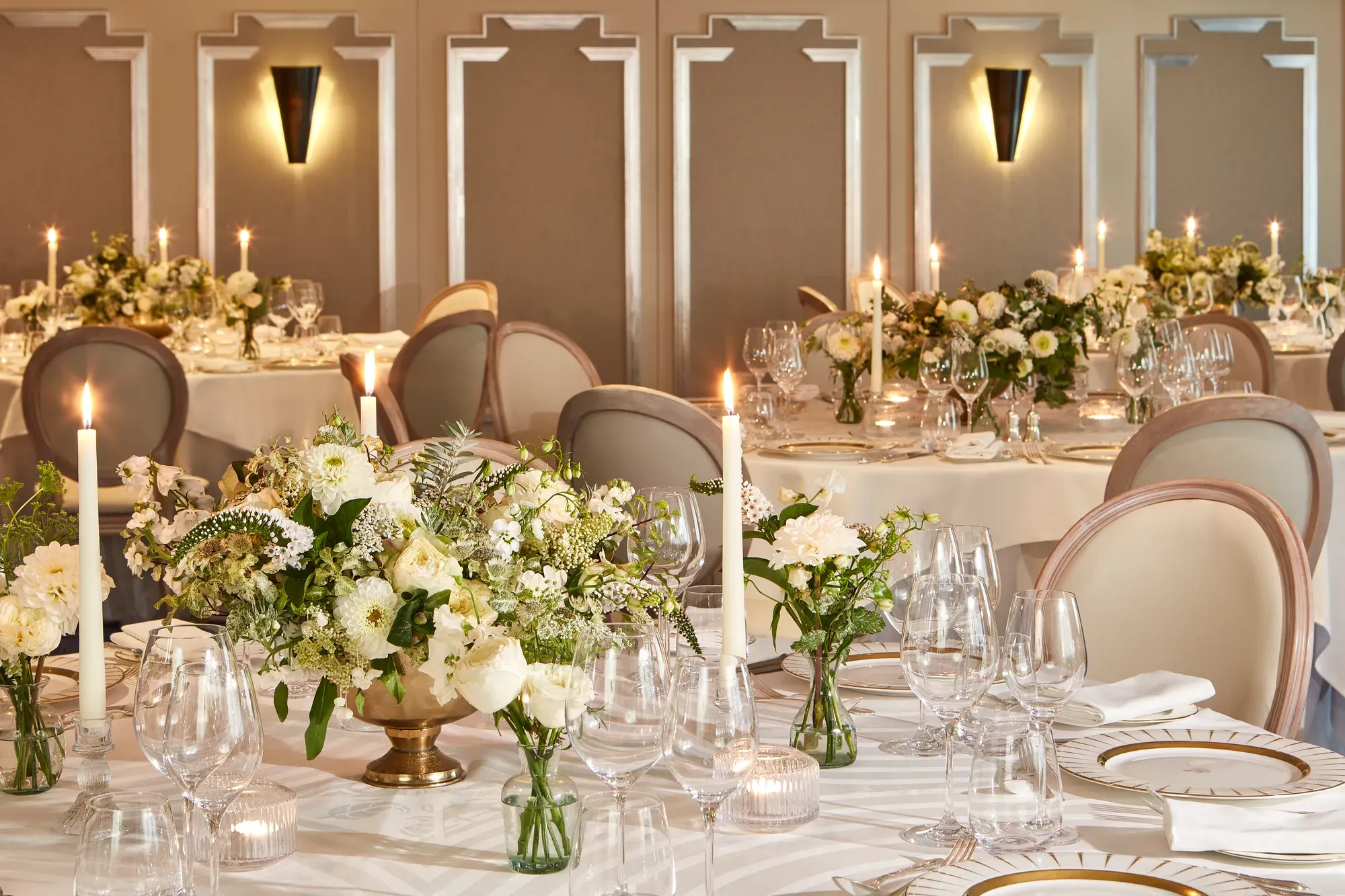 Elegant wedding reception setup in a softly lit dining room, featuring round tables dressed in white linen, gold-rimmed plates, crystal glassware, and floral centrepieces of white roses and greenery surrounded by tall lit candles.