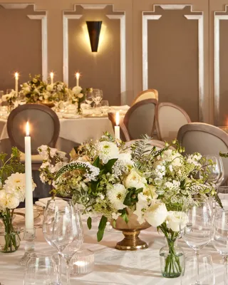 Elegant wedding reception setup in a softly lit dining room, featuring round tables dressed in white linen, gold-rimmed plates, crystal glassware, and floral centrepieces of white roses and greenery surrounded by tall lit candles.