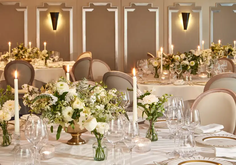 Elegant wedding reception setup in a softly lit dining room, featuring round tables dressed in white linen, gold-rimmed plates, crystal glassware, and floral centrepieces of white roses and greenery surrounded by tall lit candles.