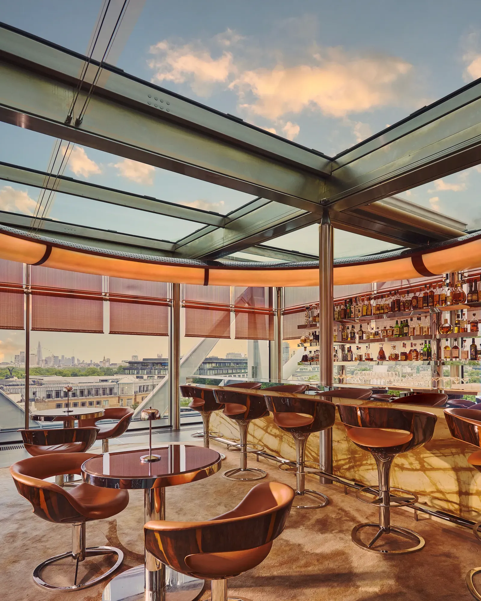 Contemporary rooftop bar with marble counter and leather stools, overlooking a city skyline through floor-to-ceiling windows.