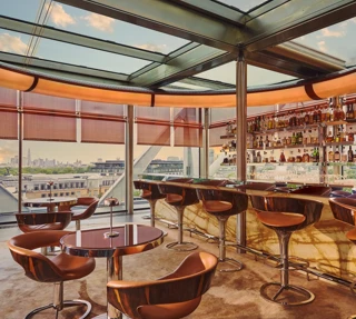 Contemporary rooftop bar with marble counter and leather stools, overlooking a city skyline through floor-to-ceiling windows.