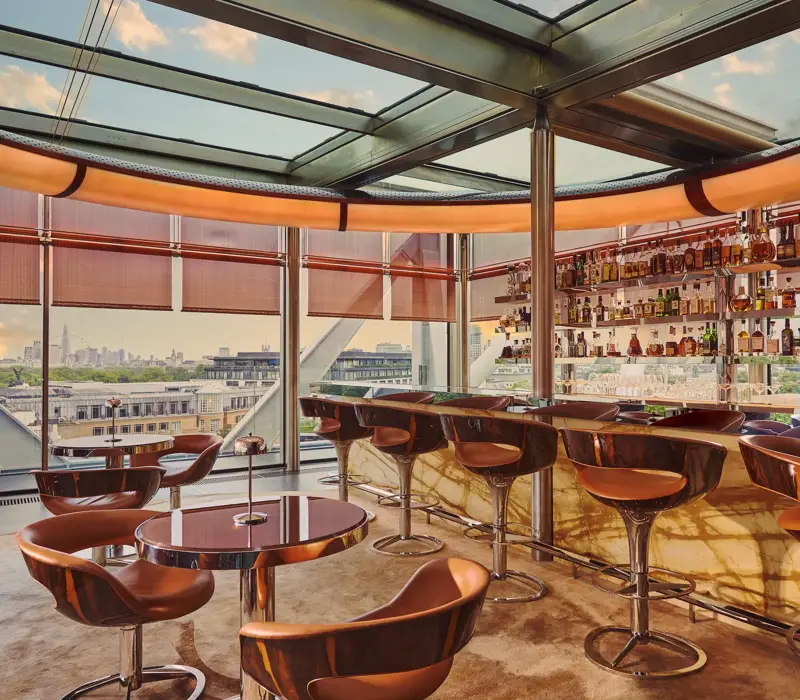 Contemporary rooftop bar with marble counter and leather stools, overlooking a city skyline through floor-to-ceiling windows.