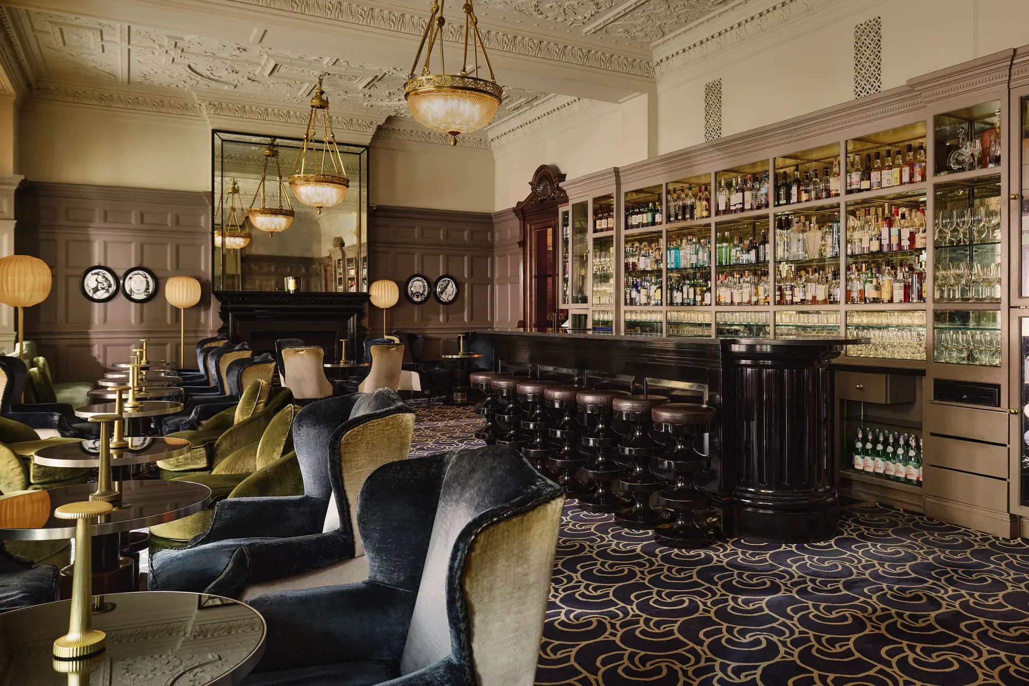 Elegant lounge bar with velvet armchairs, a long dark counter, and shelves of bottles under ornate chandeliers.