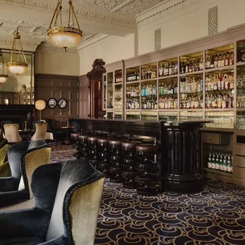 Elegant lounge bar with velvet armchairs, a long dark counter, and shelves of bottles under ornate chandeliers.