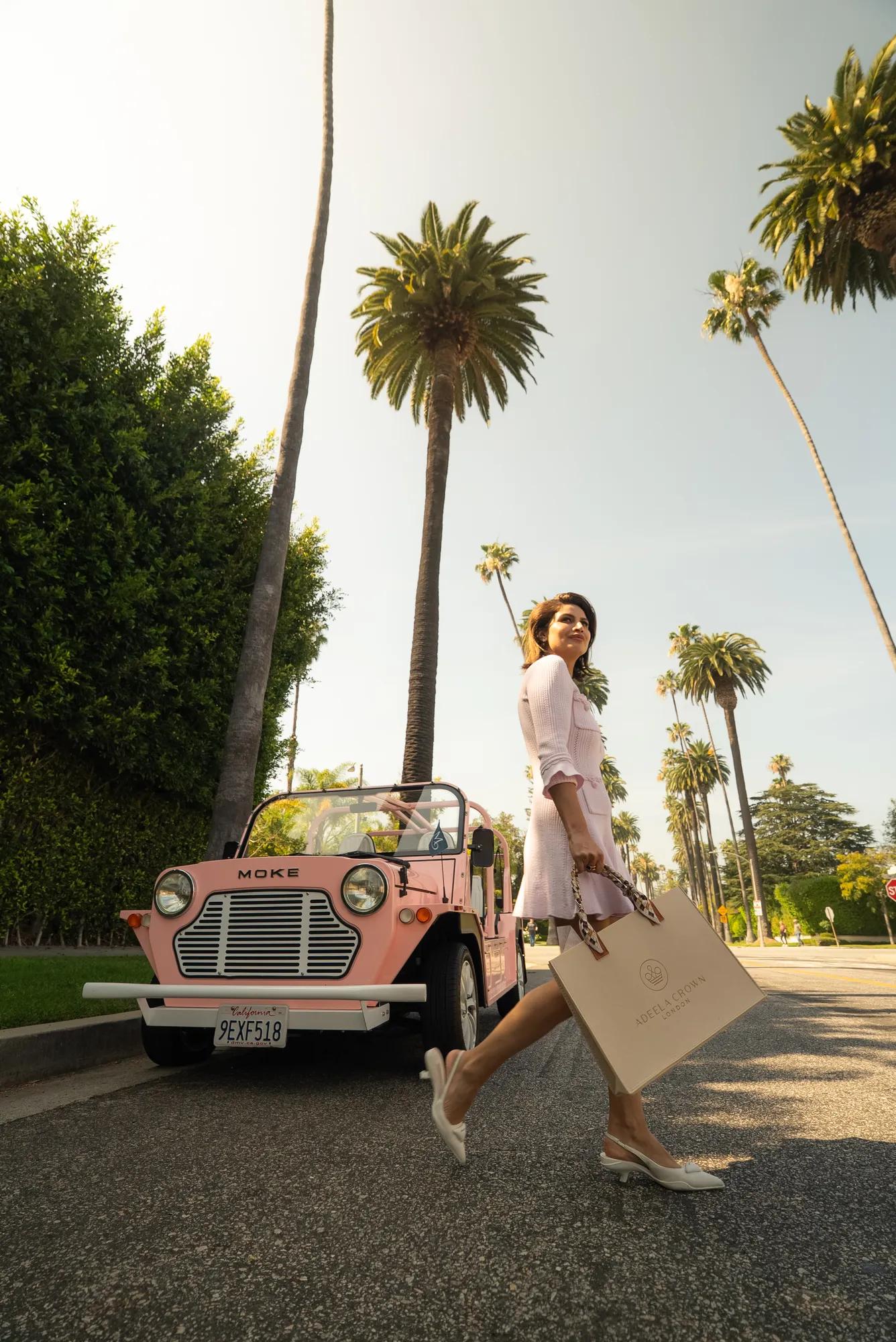 Person walking past a pink vintage car on a palm-lined street, holding a shopping bag, in a bright sunny outdoor setting.