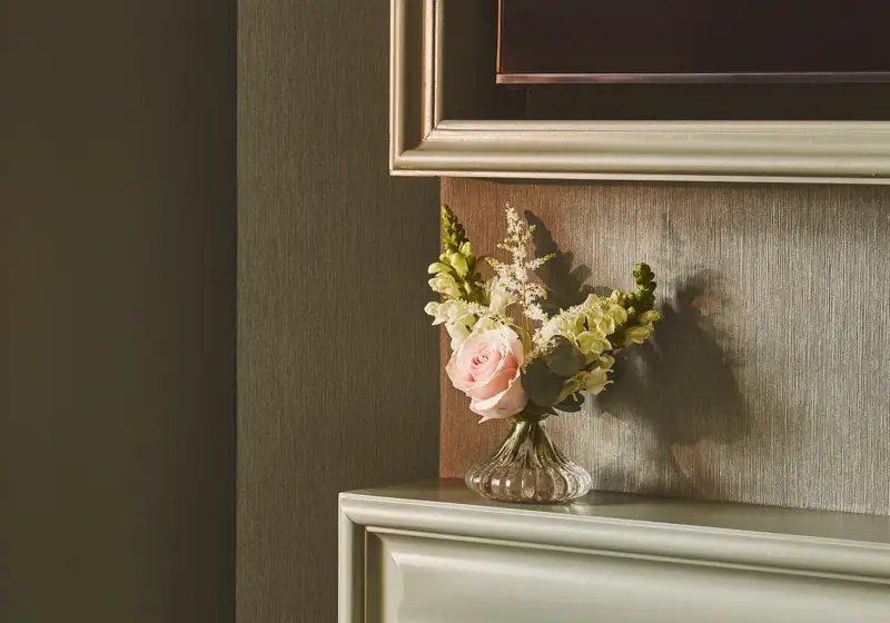 Close-up of a pale ornate fireplace with patterned tiles and a small vase of pink and white flowers on the mantel.