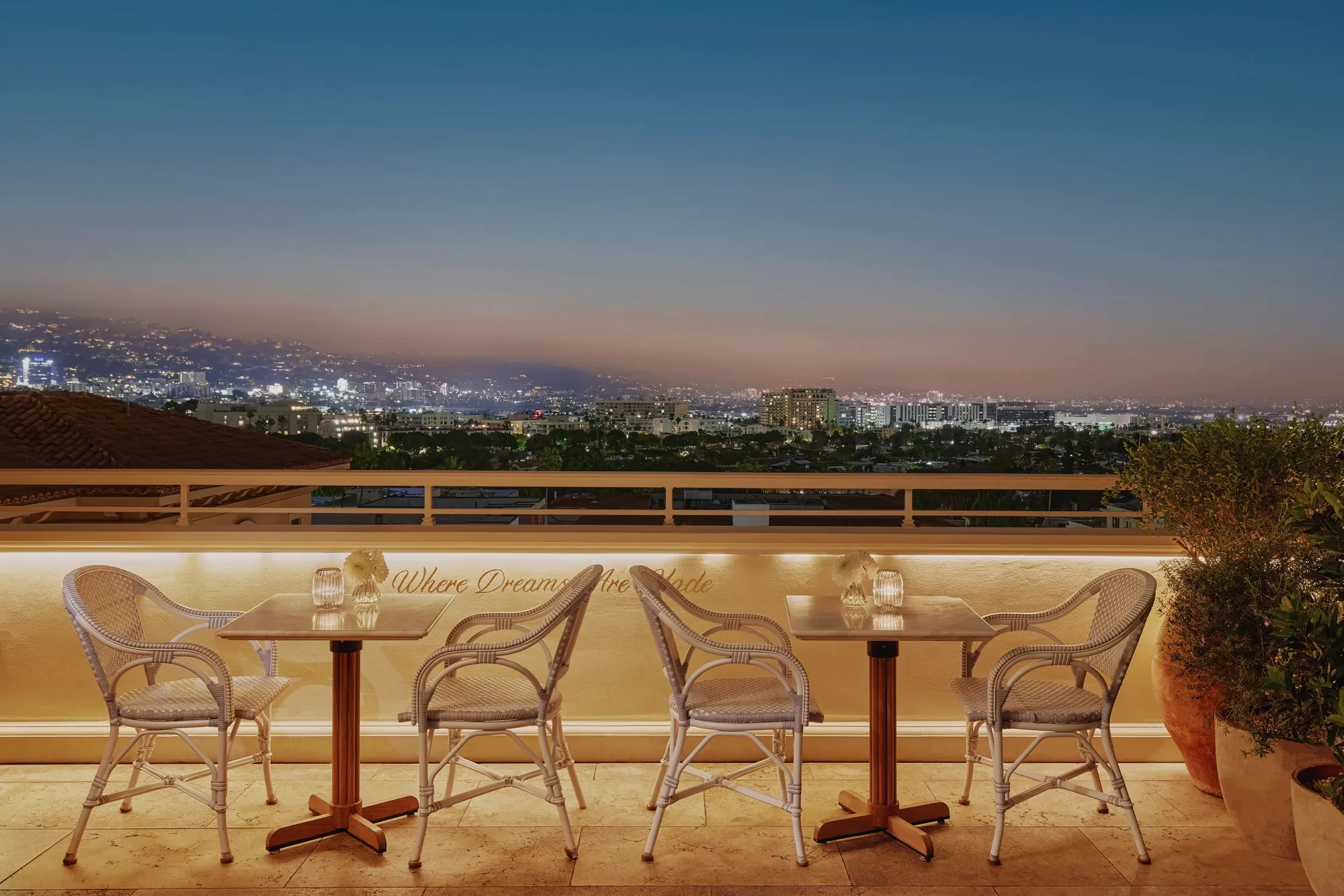 Evening terrace with café tables and wicker chairs overlooking Beverly Hills and the Hollywood Hills at dusk.