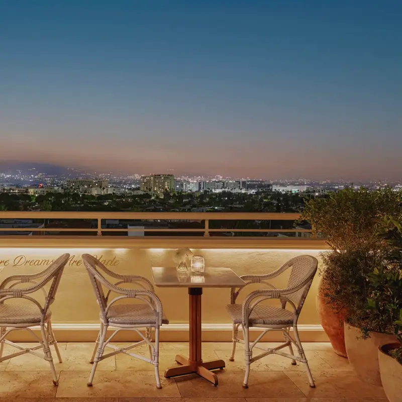 Evening terrace with café tables and wicker chairs overlooking Beverly Hills and the Hollywood Hills at dusk.