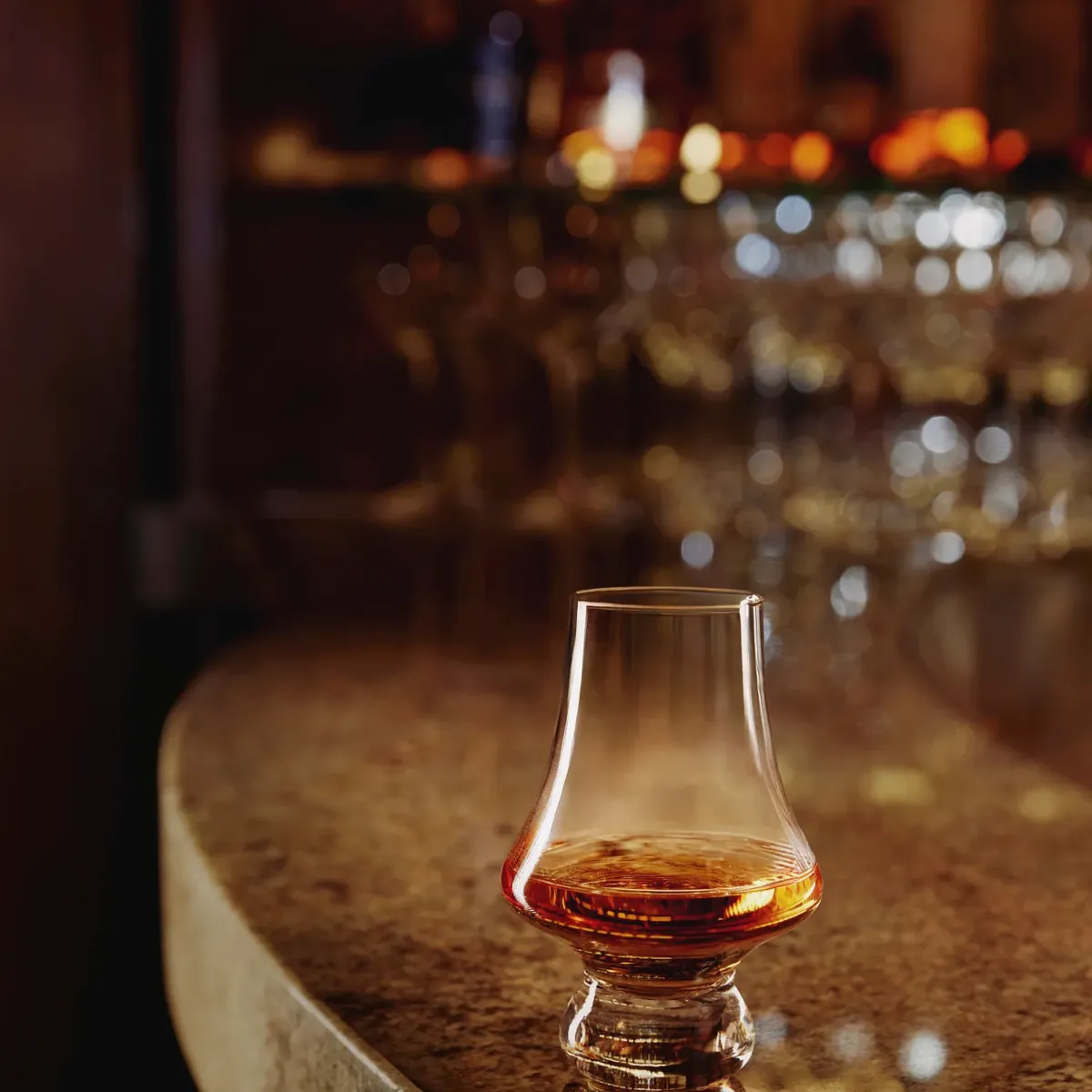 Glencairn glass of whisky on a polished granite bar with warm, out-of-focus bar lights.