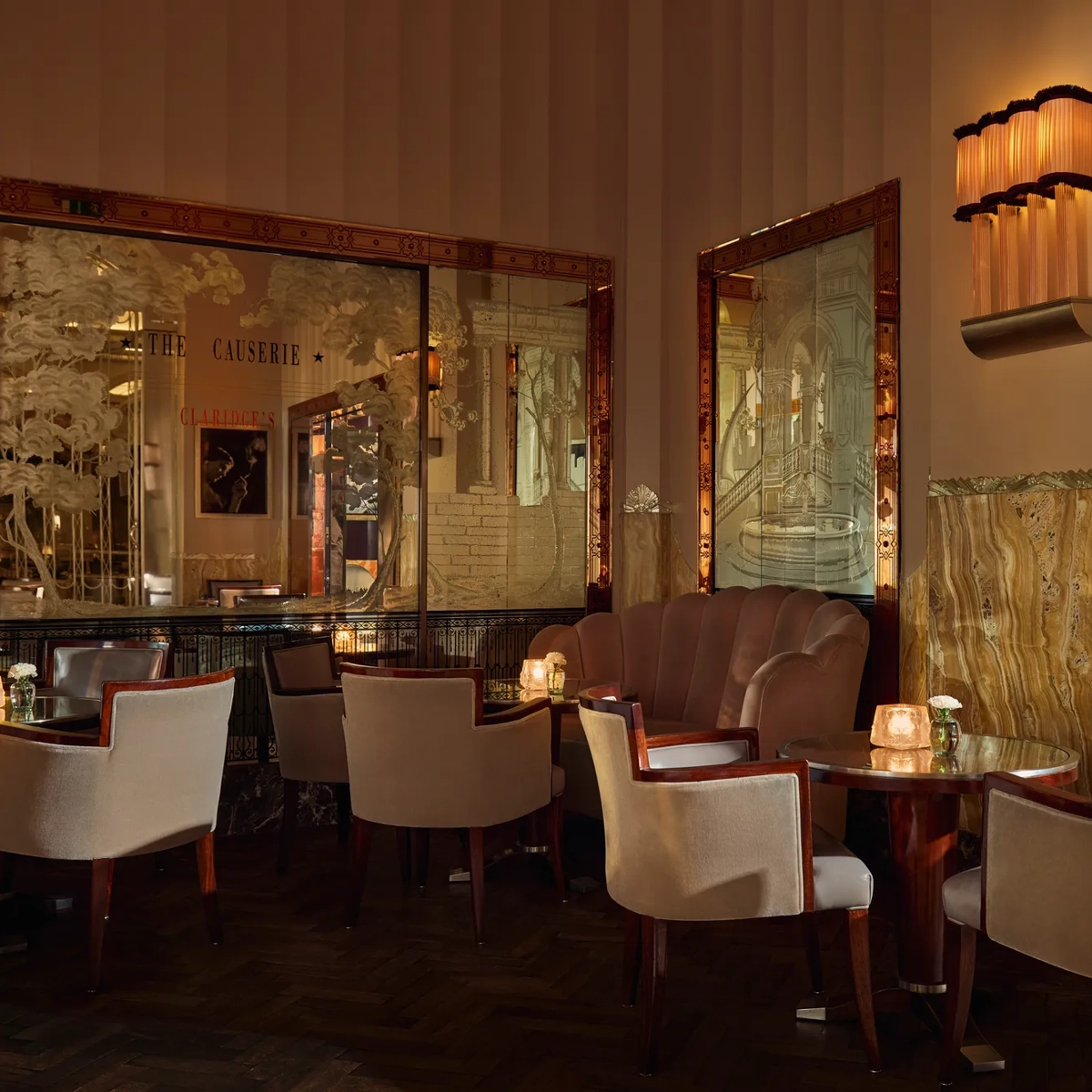 Elegant art deco lounge with soft lighting, marble walls, etched mirrors, and round tables surrounded by cream armchairs and a pink velvet banquette, creating an intimate, sophisticated atmosphere.