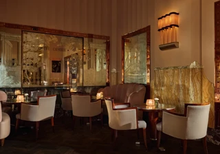 Elegant art deco lounge with soft lighting, marble walls, etched mirrors, and round tables surrounded by cream armchairs and a pink velvet banquette, creating an intimate, sophisticated atmosphere.