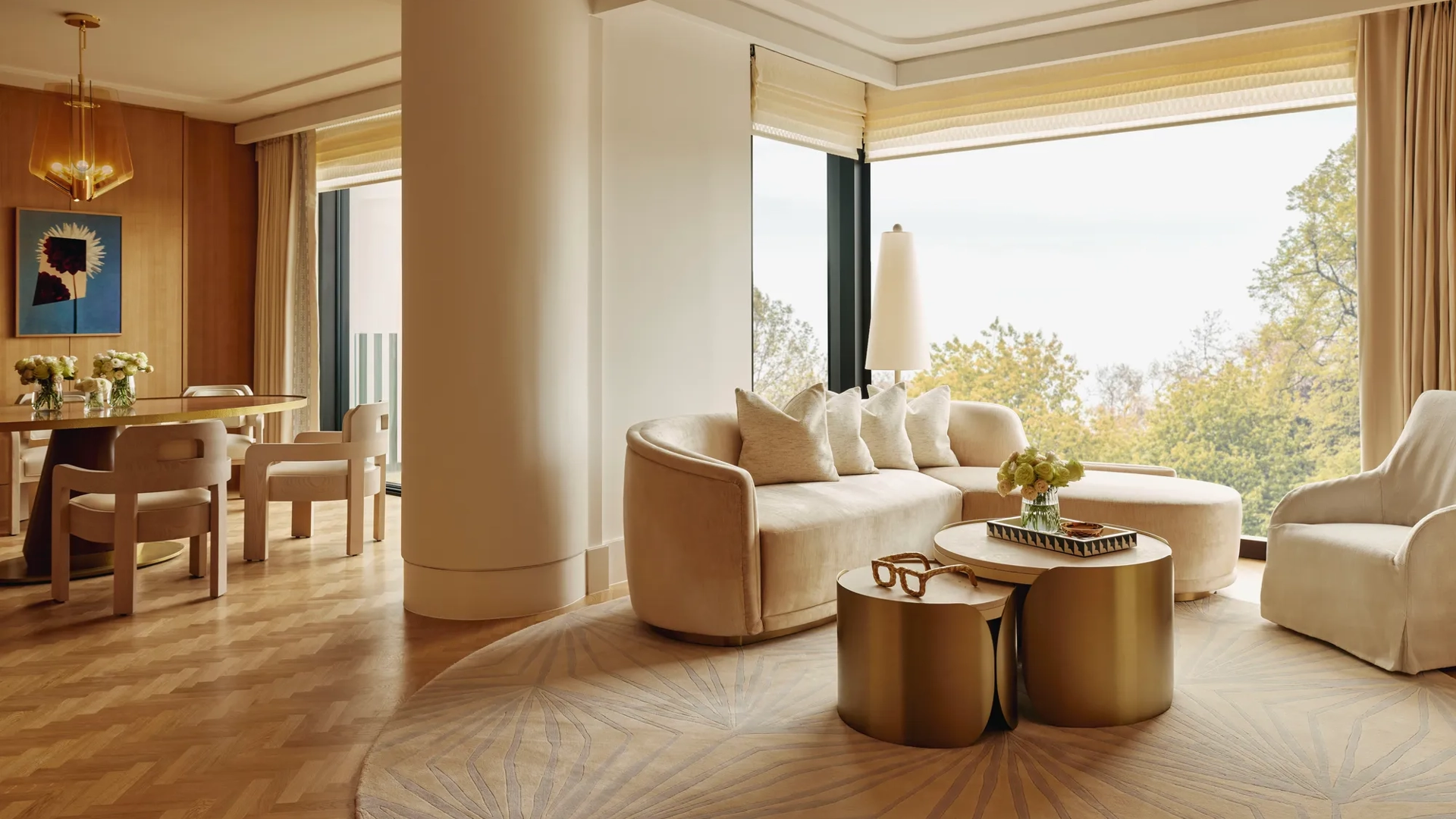 Elegant suite with curved cream sofa, gold tables, and dining area, featuring large windows overlooking Hyde Park treetops.