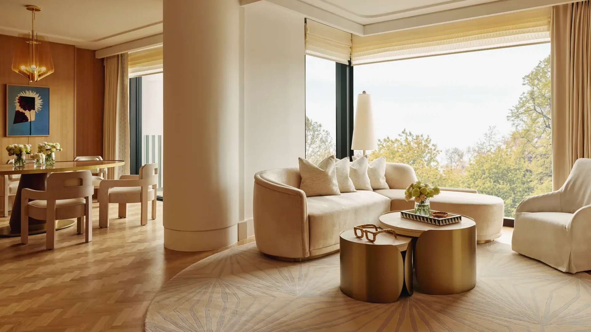 Elegant suite with curved cream sofa, gold tables, and dining area, featuring large windows overlooking Hyde Park treetops.