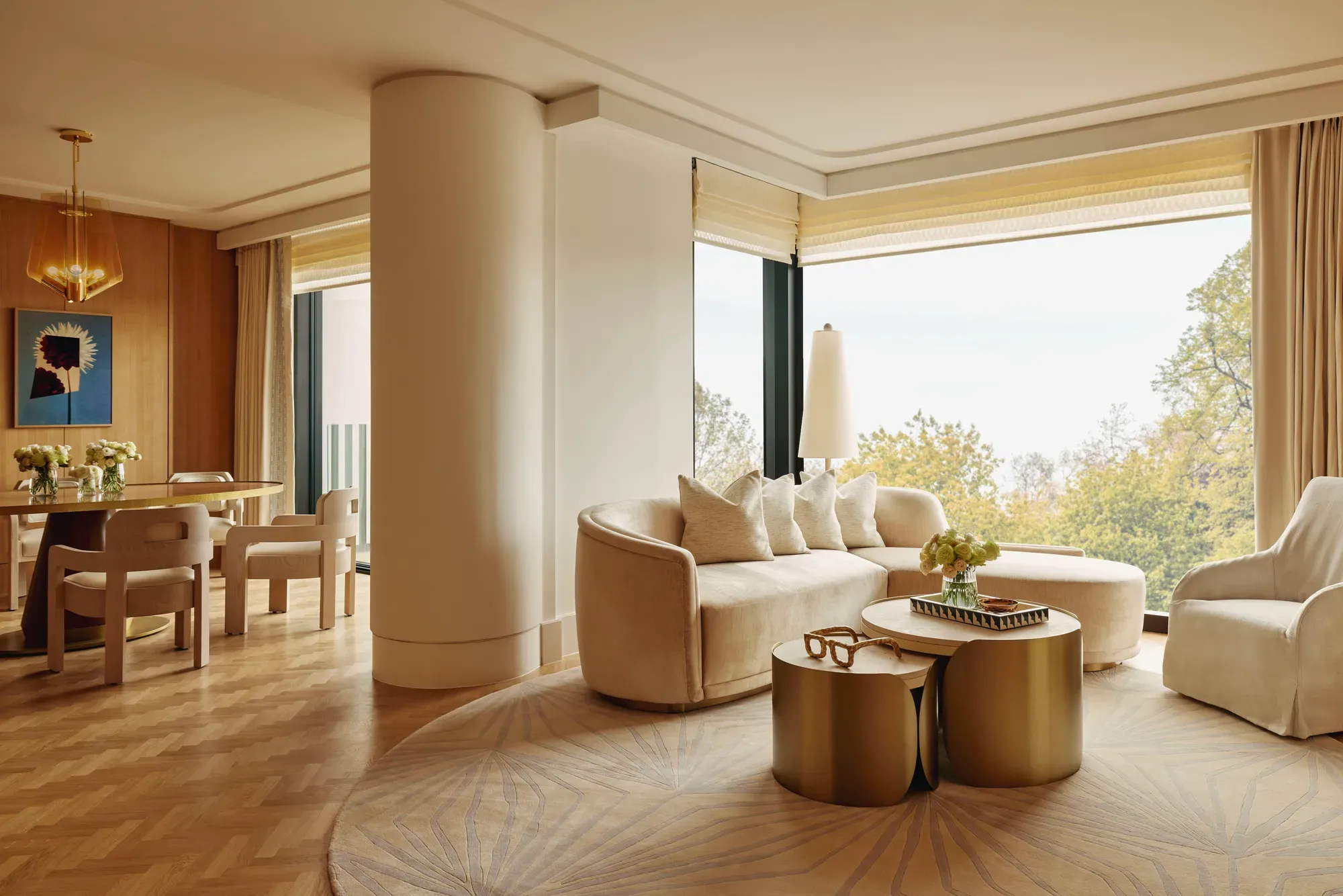 Elegant suite with curved cream sofa, gold tables, and dining area, featuring large windows overlooking Hyde Park treetops.