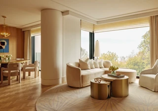 Elegant suite with curved cream sofa, gold tables, and dining area, featuring large windows overlooking Hyde Park treetops.