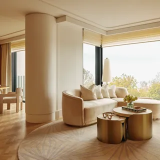 Elegant suite with curved cream sofa, gold tables, and dining area, featuring large windows overlooking Hyde Park treetops.
