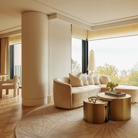 Elegant suite with curved cream sofa, gold tables, and dining area, featuring large windows overlooking Hyde Park treetops.