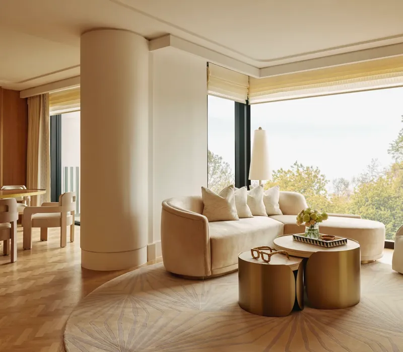 Elegant suite with curved cream sofa, gold tables, and dining area, featuring large windows overlooking Hyde Park treetops.
