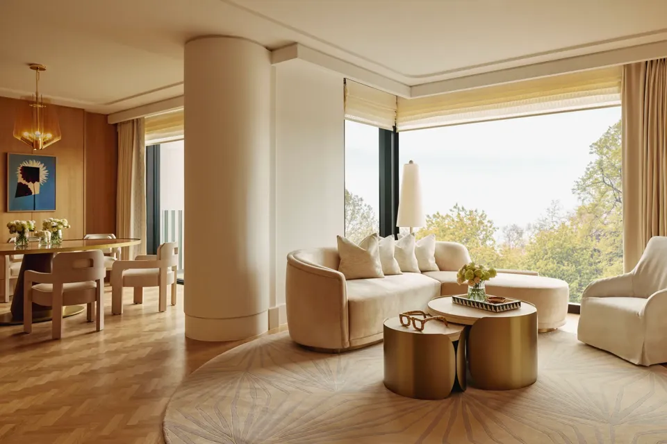 Elegant suite with curved cream sofa, gold tables, and dining area, featuring large windows overlooking Hyde Park treetops.