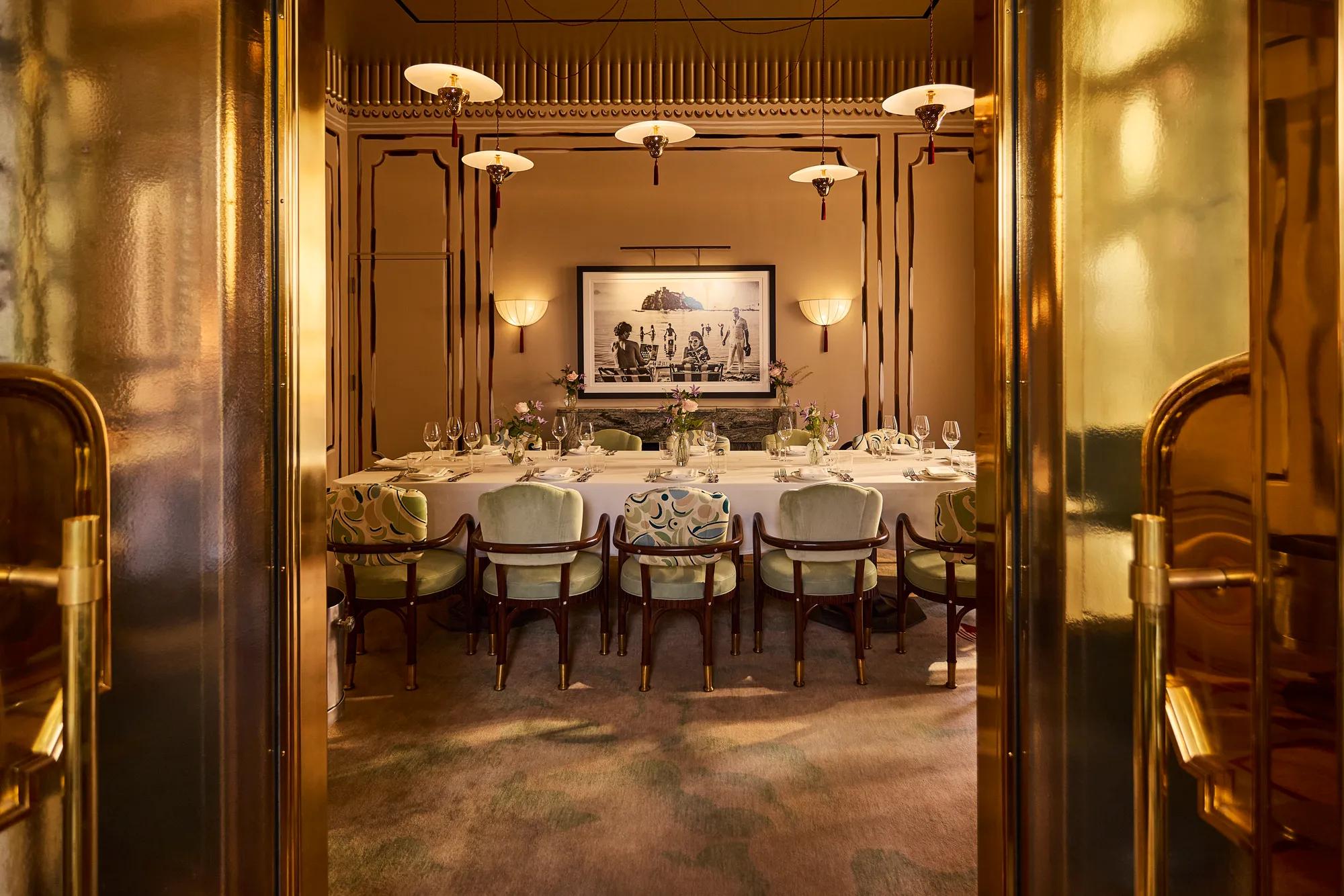 Private dining room viewed through bronze doors, featuring a long dressed table, upholstered chairs and warm Art Deco details.