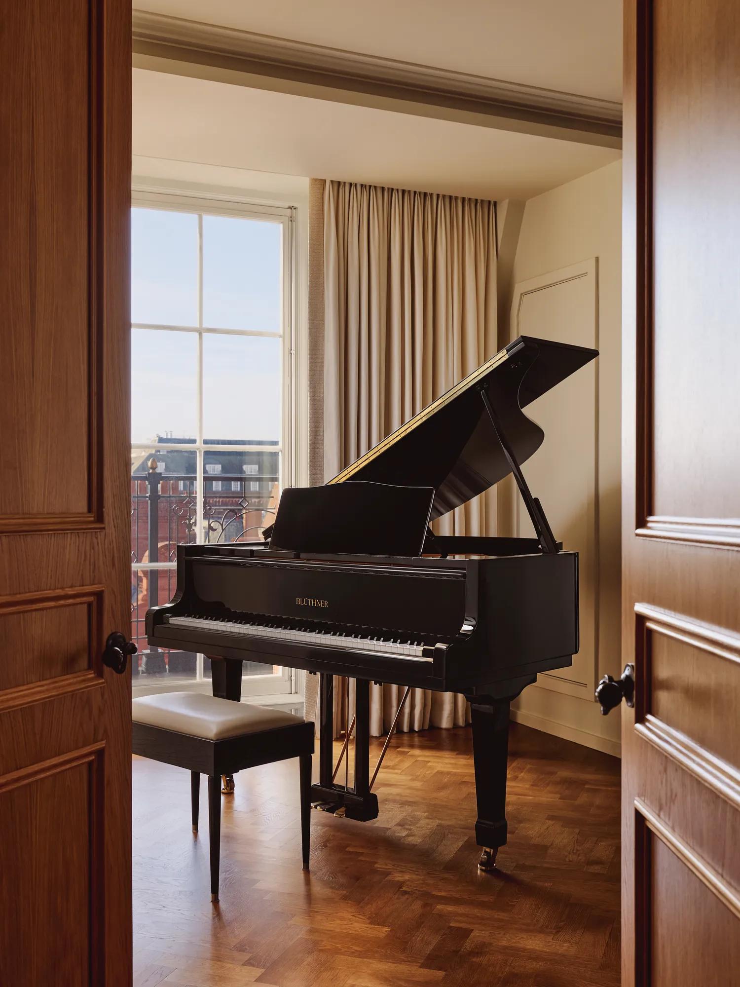 Elegant music room with a black grand piano and matching bench positioned beside a large window with cream curtains, overlooking red-brick buildings.