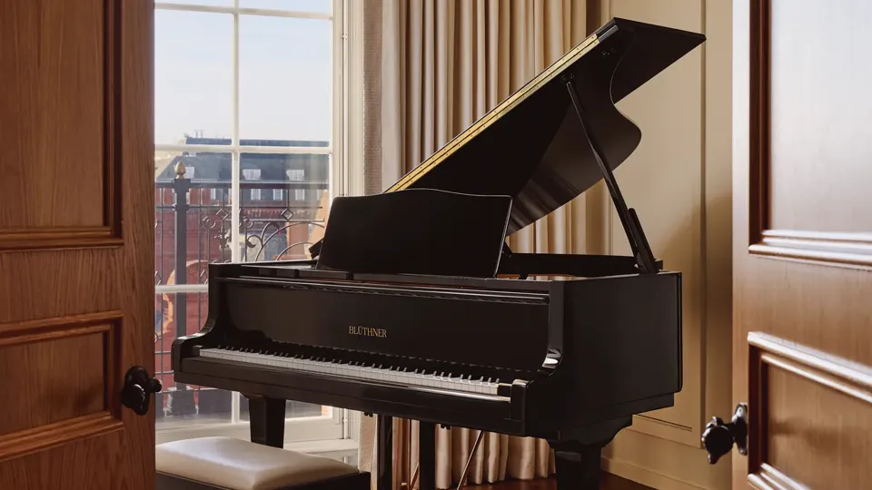 Elegant music room with a black grand piano and matching bench positioned beside a large window with cream curtains, overlooking red-brick buildings.