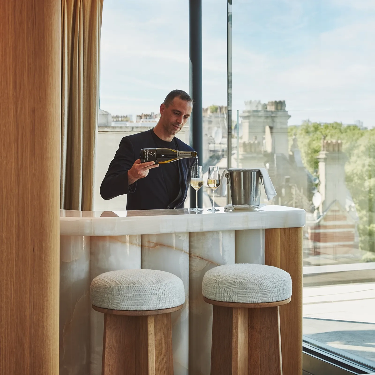 Man pouring wine at a marble in-room bar with stools, beside a large window overlooking city rooftops.
