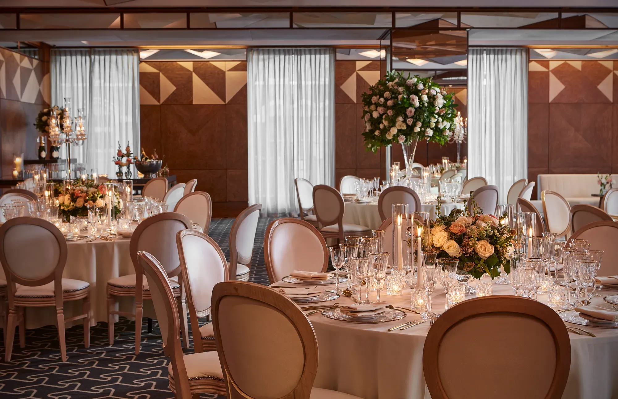 Ballroom with round tables, white chairs, and floral centrepieces lit by candles for an elegant wedding reception.