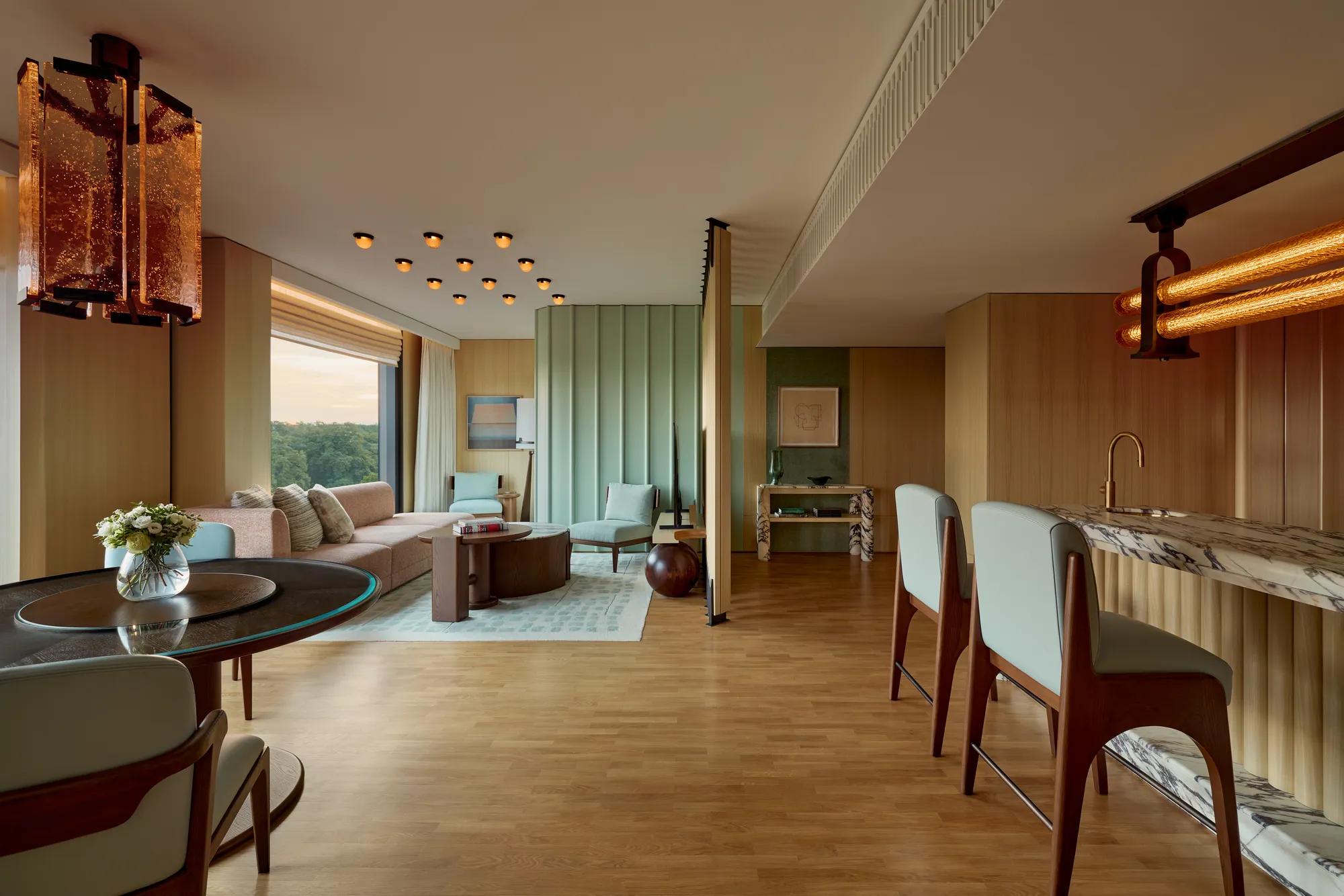 Spacious modern suite with marble bar, dining table, seating area, pastel green wall panels, large windows, and contemporary lighting overlooking greenery.