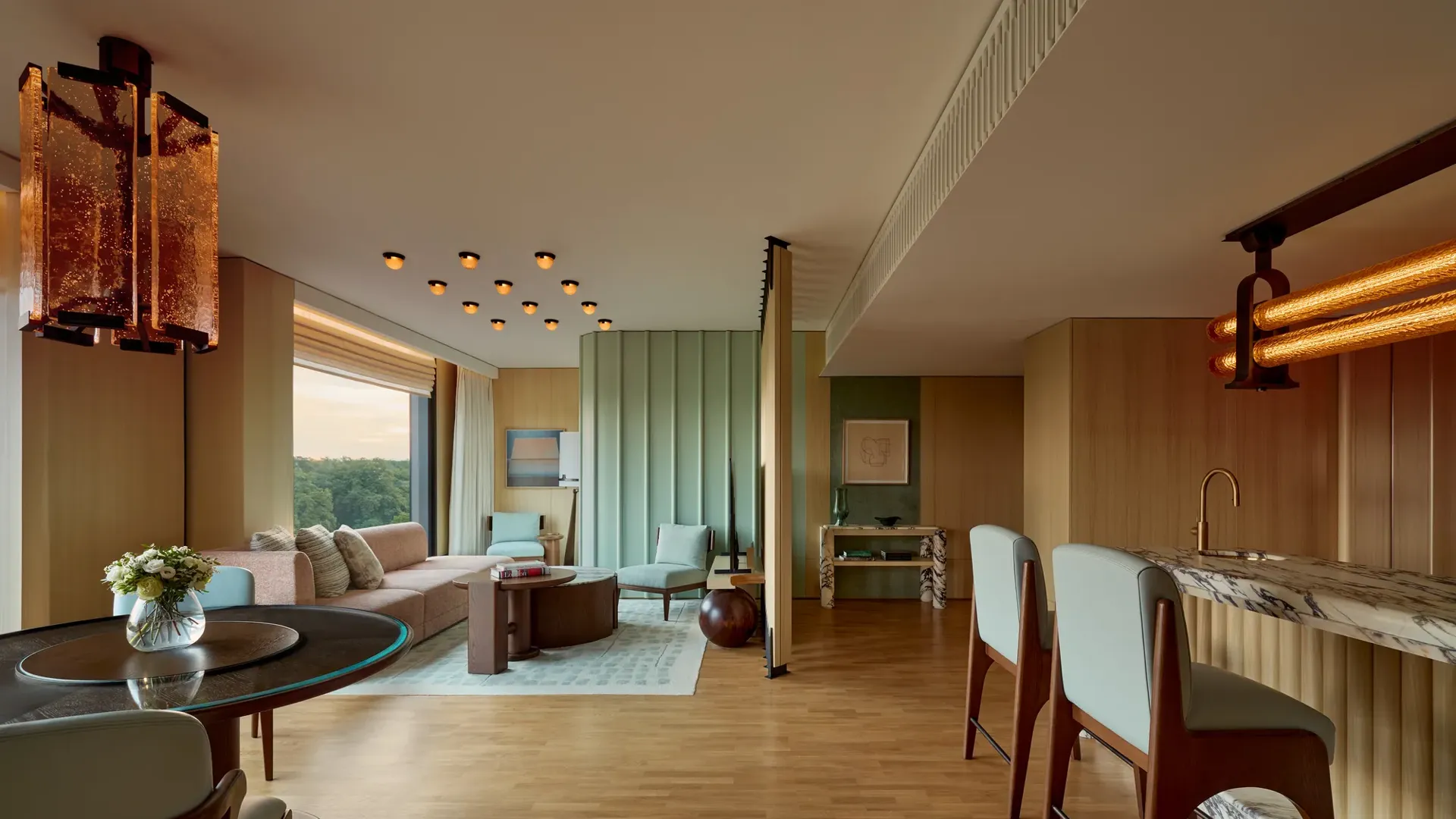 Spacious modern suite with marble bar, dining table, seating area, pastel green wall panels, large windows, and contemporary lighting overlooking greenery.