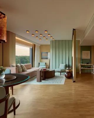 Spacious modern suite with marble bar, dining table, seating area, pastel green wall panels, large windows, and contemporary lighting overlooking greenery.