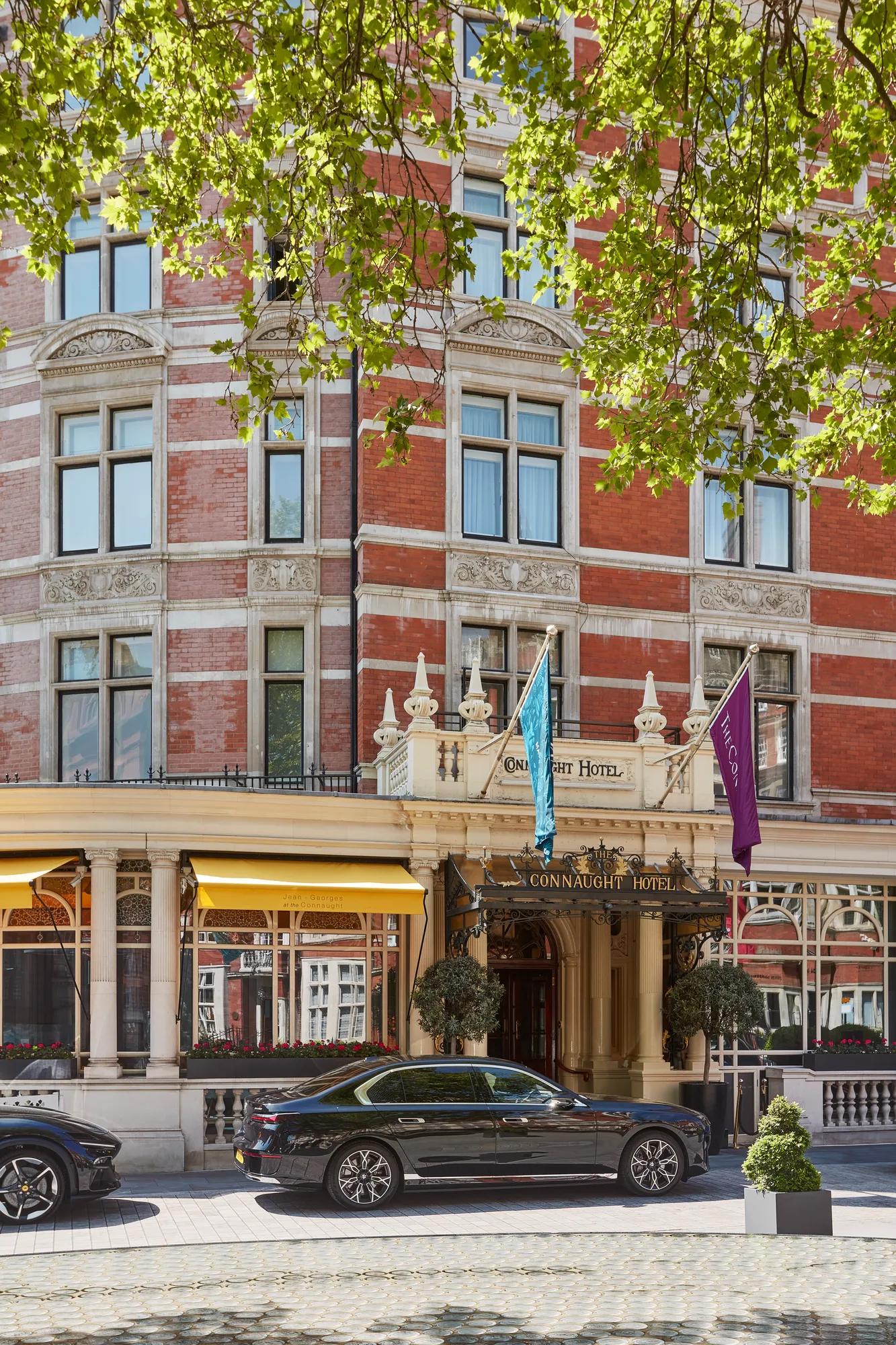 Red-brick hotel exterior with ornate entrance, flags and yellow awnings, framed by leafy branches, with a black car parked at the front.