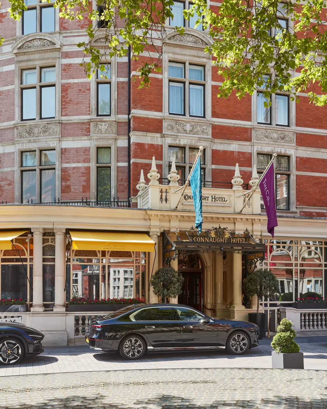 Red-brick hotel exterior with ornate entrance, flags and yellow awnings, framed by leafy branches, with a black car parked at the front.
