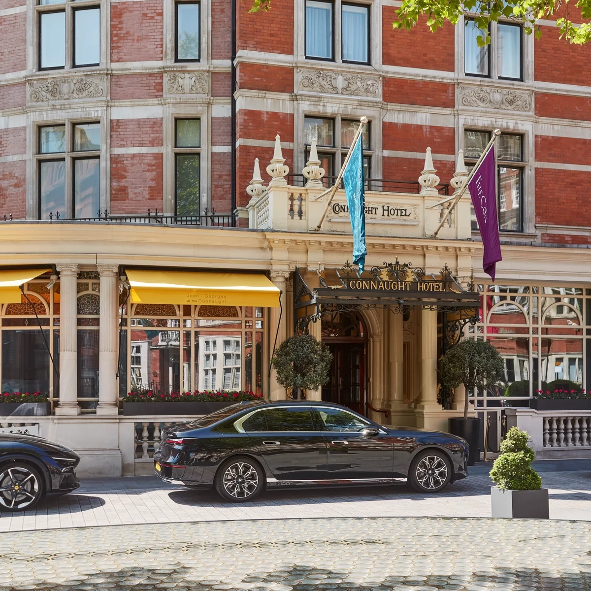 Red-brick hotel exterior with ornate entrance, flags and yellow awnings, framed by leafy branches, with a black car parked at the front.