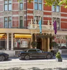 Red-brick hotel exterior with ornate entrance, flags and yellow awnings, framed by leafy branches, with a black car parked at the front.