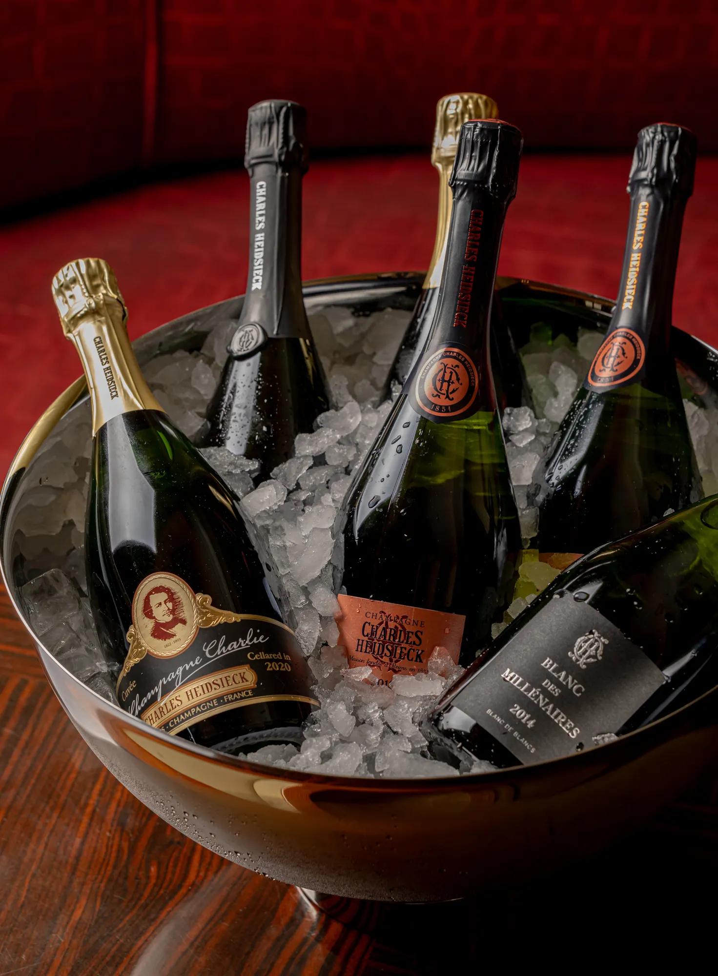 Five Champagne bottles chilling in ice-filled metal bowl on wooden table against rich red backdrop.