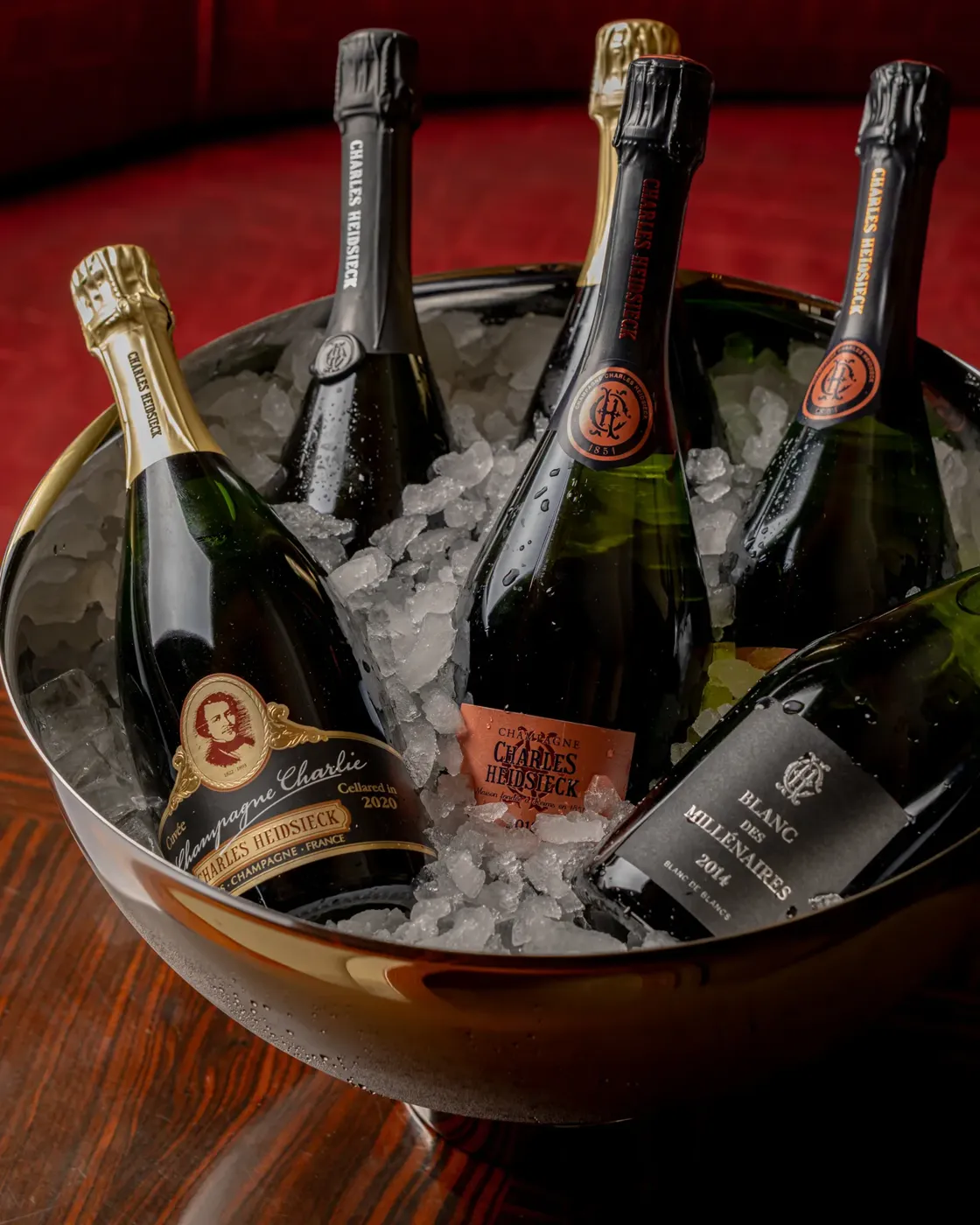 Five Champagne bottles chilling in ice-filled metal bowl on wooden table against rich red backdrop.