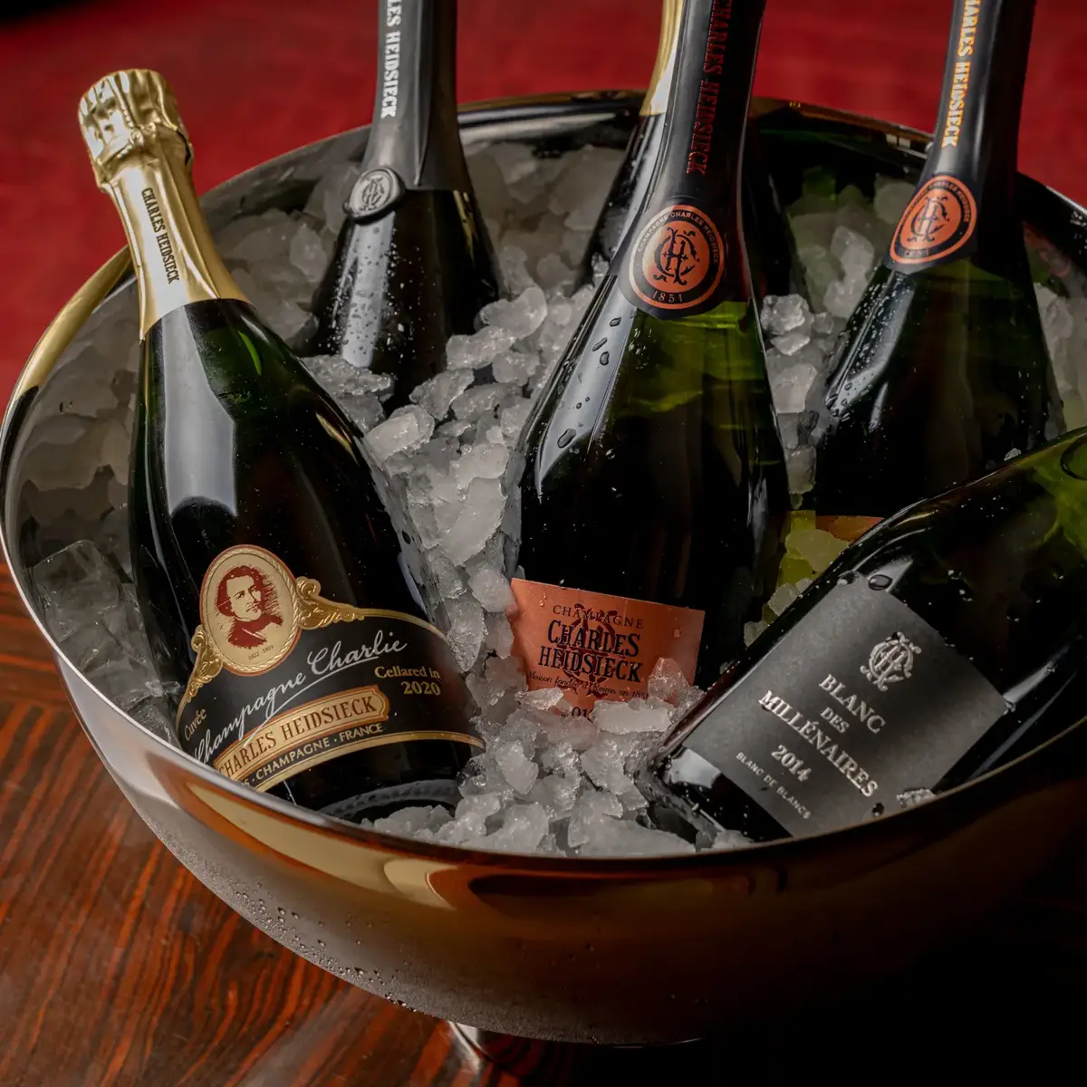 Five Champagne bottles chilling in ice-filled metal bowl on wooden table against rich red backdrop.