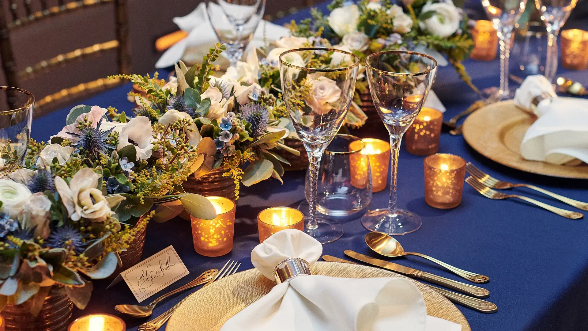 Elegant candlelit table setting with gold chargers, white napkins, and floral centerpieces on navy tablecloth.